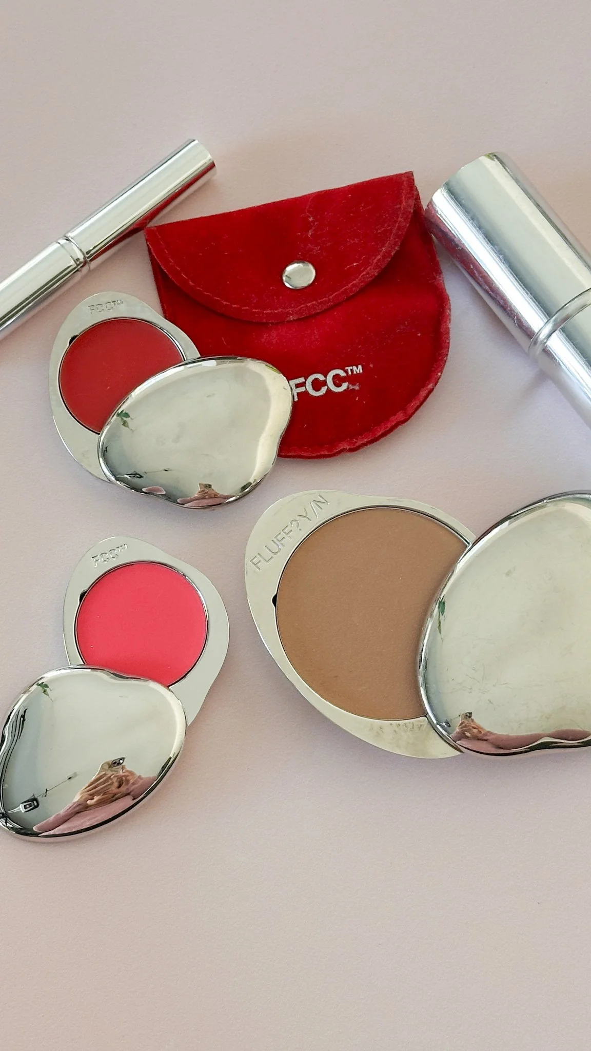 Collection of lipstick and lip balm containers, some with red and pink shades, a red velvet pouch with the FCC logo, and two silver lipsticks on a pink surface.
