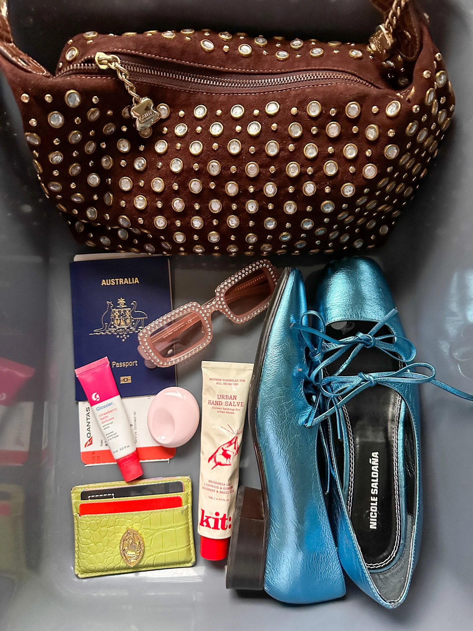 Brown studded handbag, teal shoes, pink sunglasses, Australian passport, various small toiletries, lime green wallet, and a small pink container inside a clear storage container.