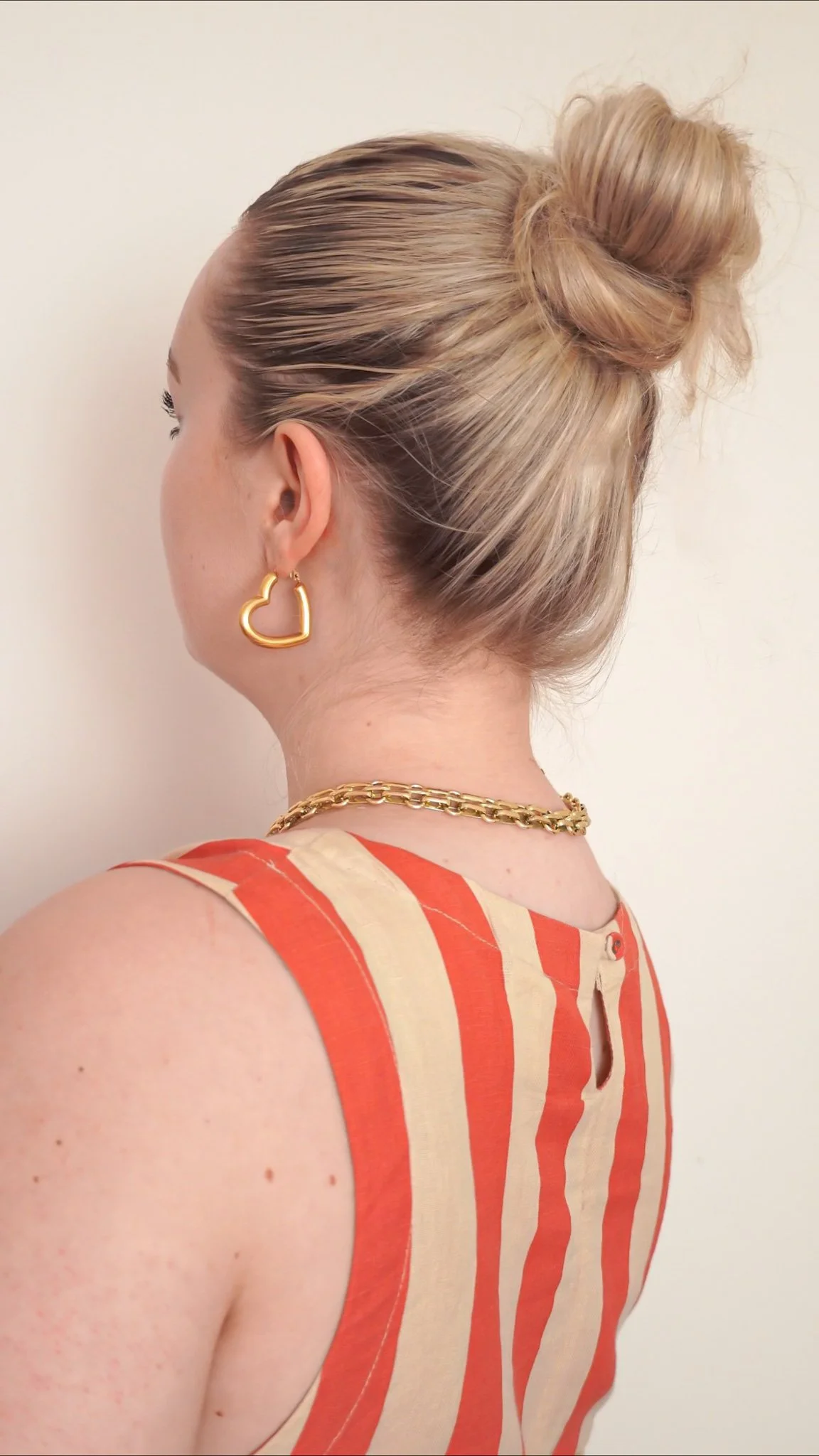 A woman with blonde hair in a bun, wearing gold heart-shaped earrings, a gold chain necklace, and a sleeveless top with orange and beige vertical stripes, standing against a plain background.