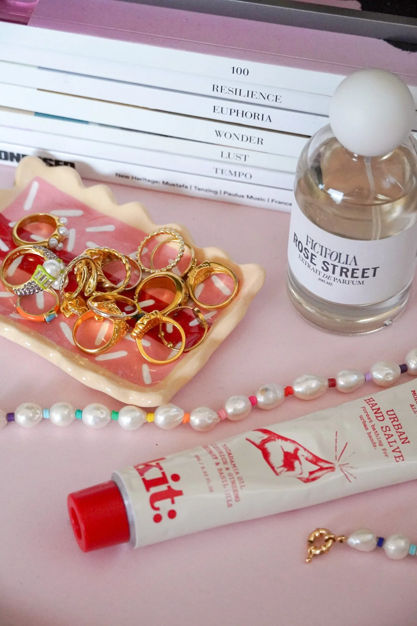 A collection of gold rings, pearl necklaces, a bottle of Ricciolia Rose Street perfume, a tube of Urban Hand Salve, and a pink plate with a pink and white pattern on a pink surface, with a book featuring words like 'Resilience,' 'Euphoria,' and 'Wond