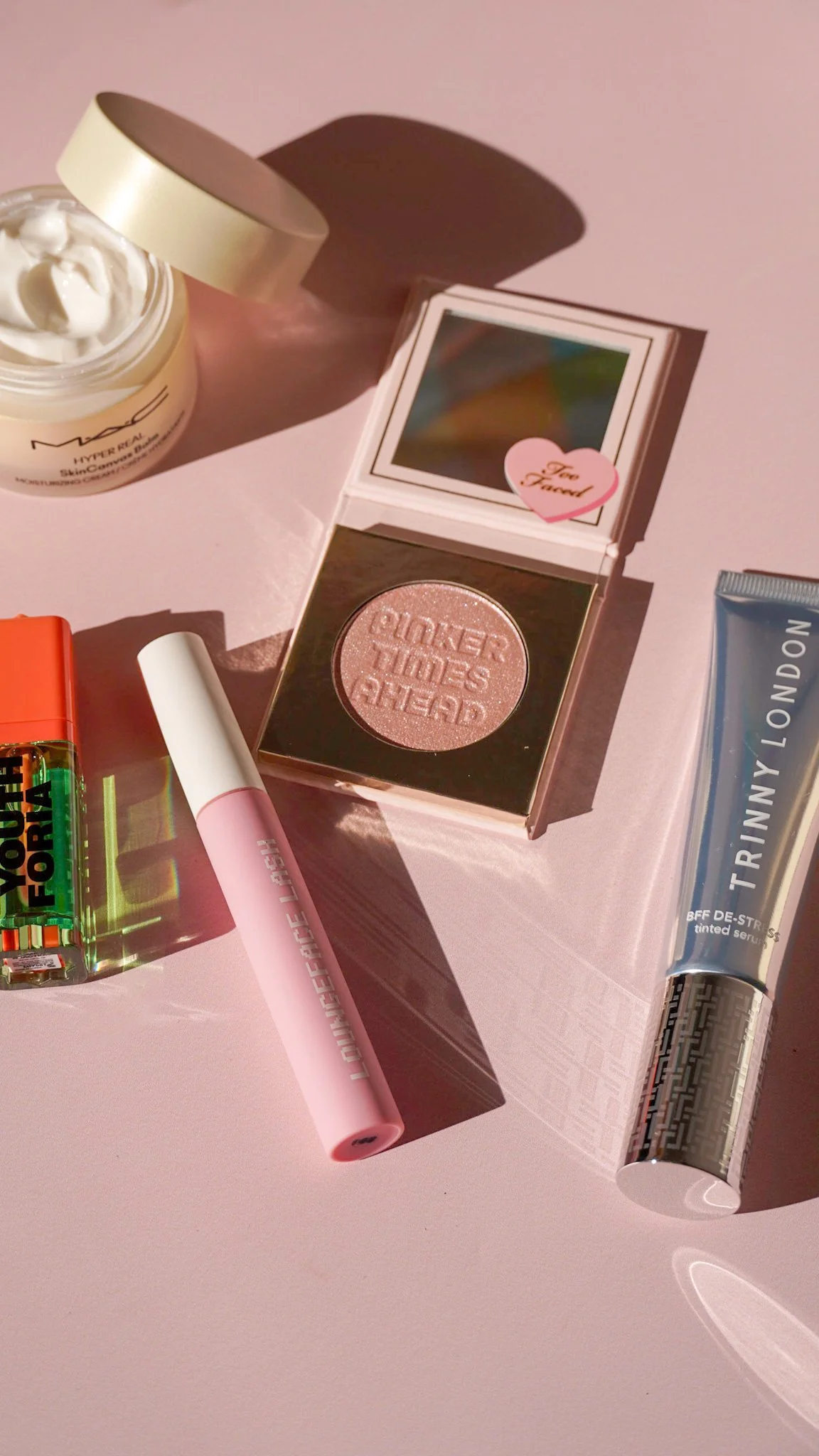 A pink surface holding beauty products, including a MAC skincare jar, a pink lip balm, a compact with a light pink pressed powder with the inscription 'PINKER TIMES AHEAD,' a tube of Trinny London tinted serum, and a colorful lipstick. A small mirror