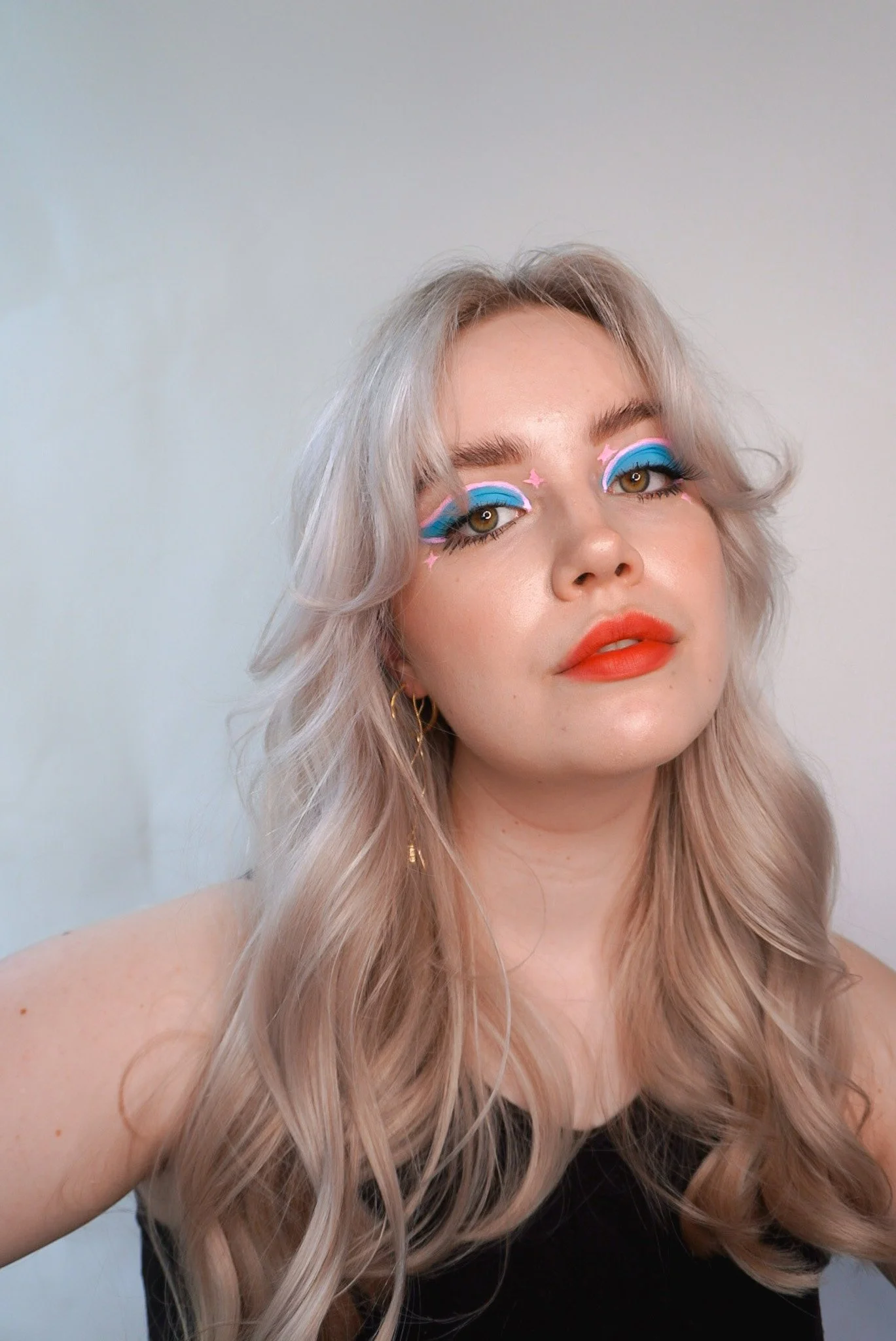 A woman with platinum blonde hair styled in loose waves wearing bold rainbow-colored eyeshadow and orange lipstick.