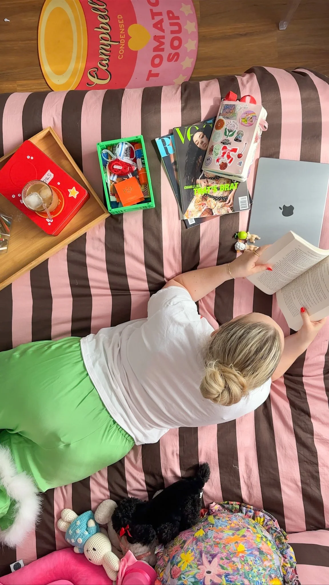 A woman lying on a striped pink and brown couch reading a book, surrounded by items including a MacBook, magazines, toys, stickers, a tray with a glass of iced coffee, and a large round Charlie Brown themed container, with a dog laying nearby.