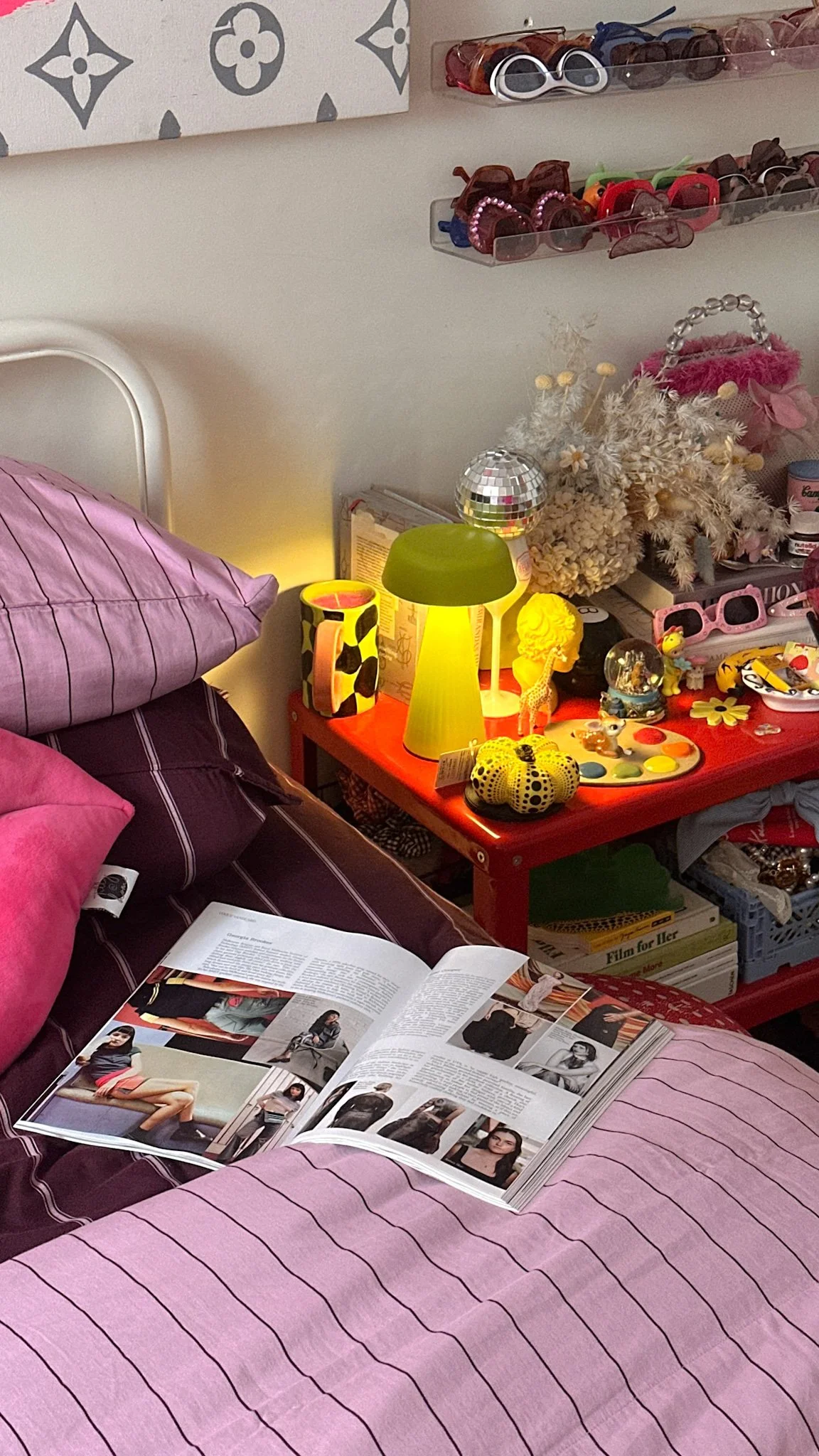 A pink-striped bed with a magazine open on it, next to a red nightstand with a green lamp, decorative items, and a cloudy white book underneath a shelf of colorful sunglasses. There are also pink pillows and a pink blanket on the bed.