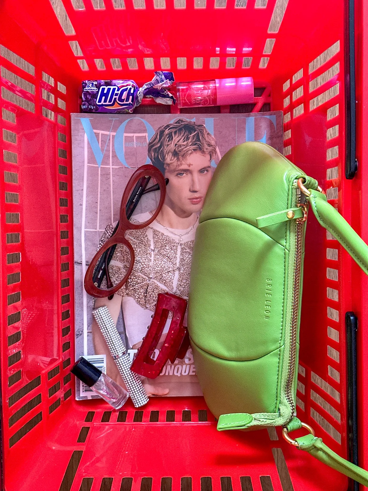 Red shopping basket containing a Vogue magazine with a woman on the cover, a pair of red sunglasses, a clear nail polish bottle, a red hair clip, a white dotted item, and a lime green purse.