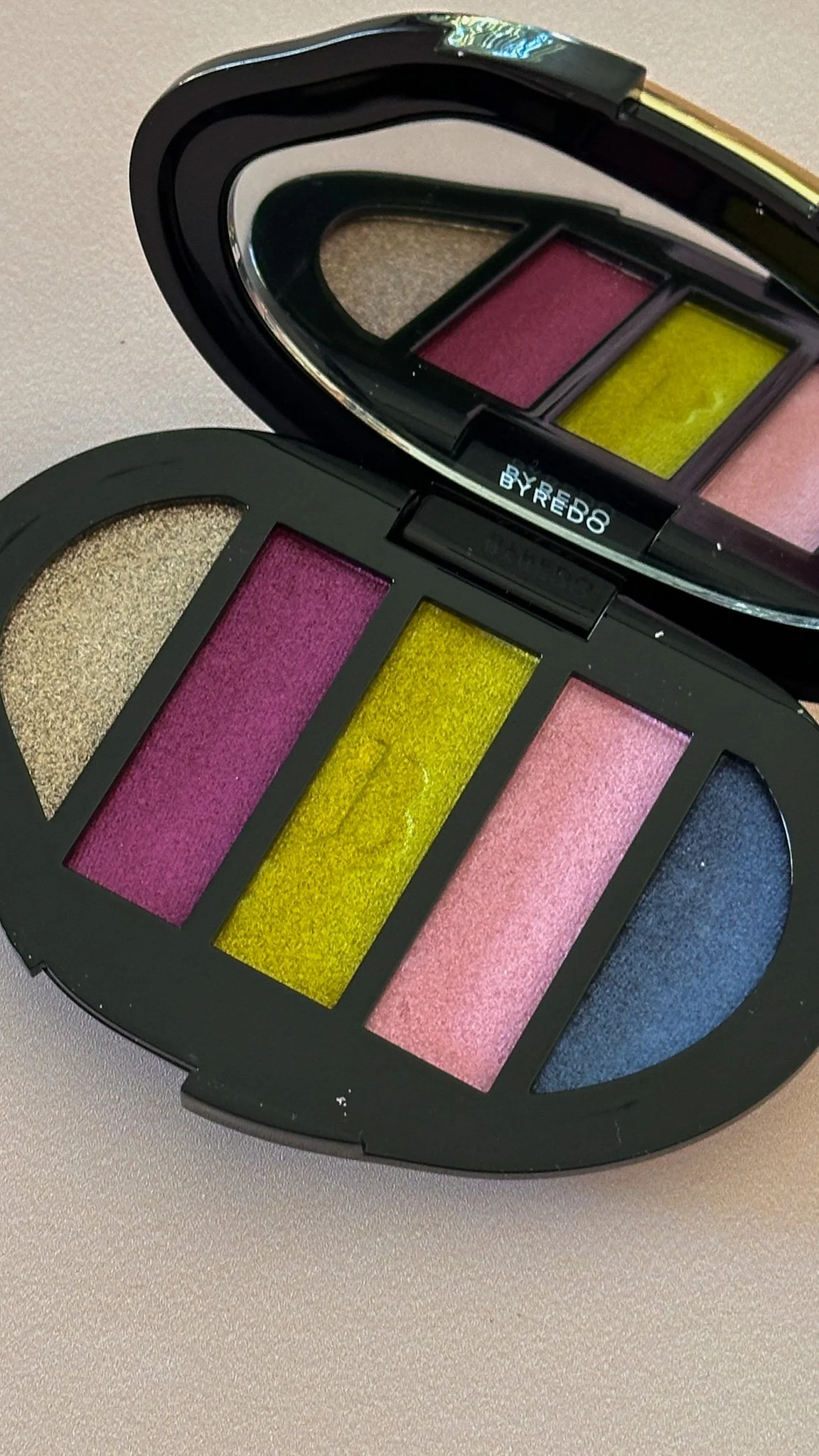 A colorful eyeshadow palette with shades of gold, purple, yellow, pink, and blue, housed in a black case with a mirror.
