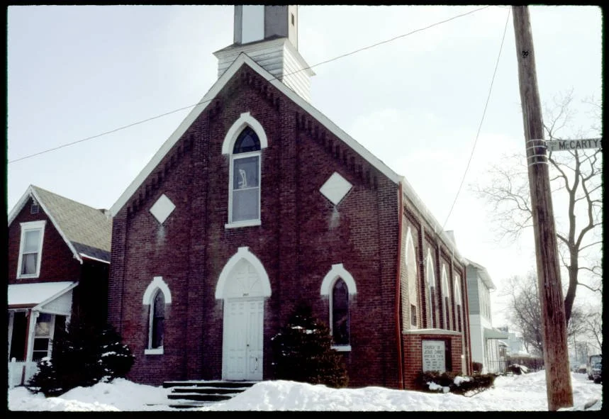 1978 photo of historic Danish Lutheran church with a white door and arched windows, surrounded by snow, with a tall steeple at the top.