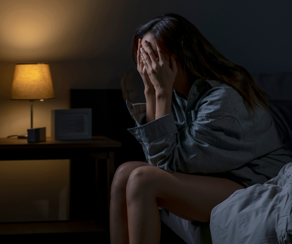 woman sitting in darkly lit bedroom on edge of bed in pajamas with her head in her hands upset