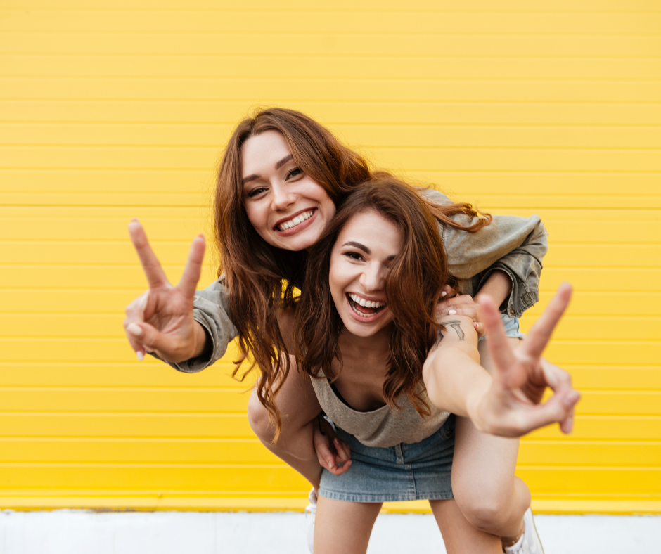 two young women with red hair laughing one on the others back holding up the peace sign and smiling in front of a bright yellow brick wall