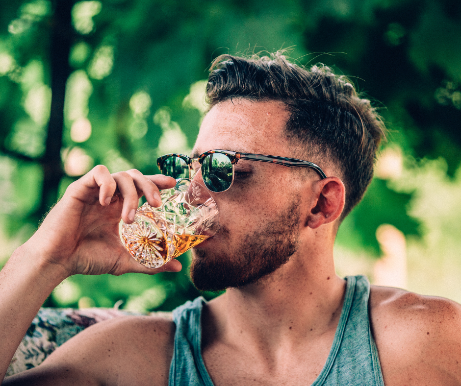 green wooded background headshot of a youn man with light brown hair and black sunglasses drinking shot of liquor