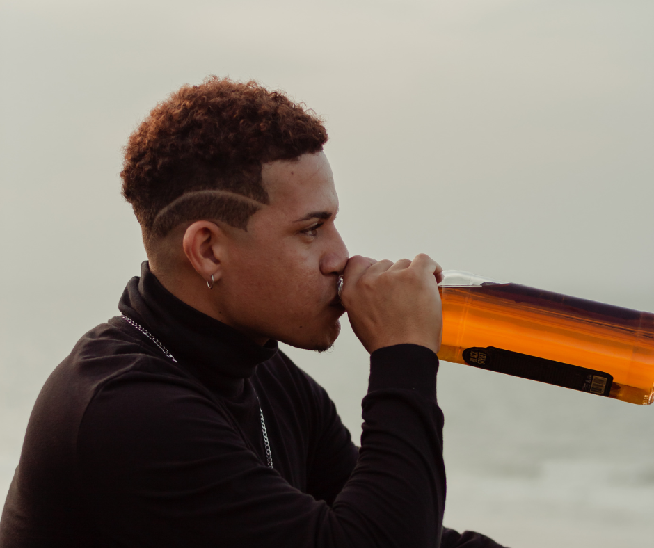 young African American male sitting down looking straight ahead holding a bottle of alcohol up to his mouth with light beige background