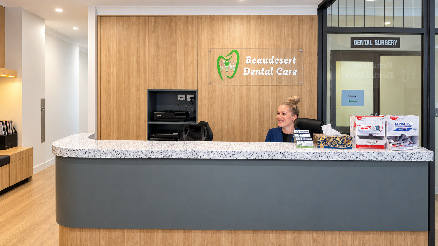 Reception area at Beaudesert Dental Care with smiling receptionist behind a speckled white countertop, dental supplies on the right, wooden wall with dental clinic sign, and a window leading to the dental surgery entrance.