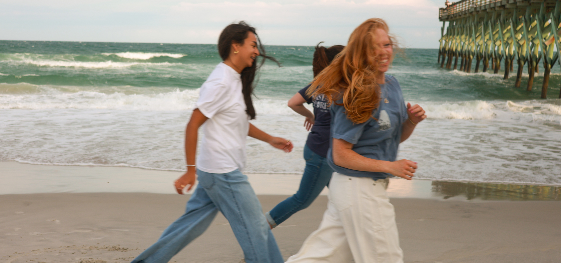 Three women running on a beach with waves and a pier in the background, smiling and enjoying the day.