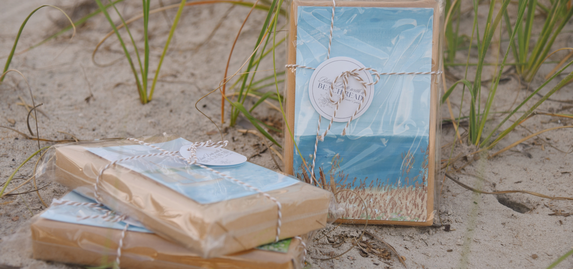 Three wrapped beach-themed gifts on sand among green beach grass, with one gift standing and two lying flat.