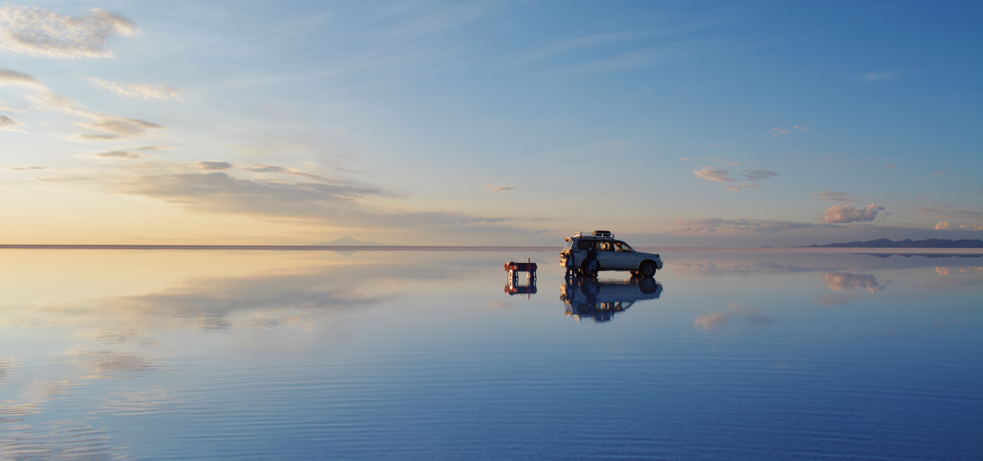 A white SUV parked near water with a small table and chairs, reflecting in the still water, under a partly cloudy sky at sunset or sunrise.