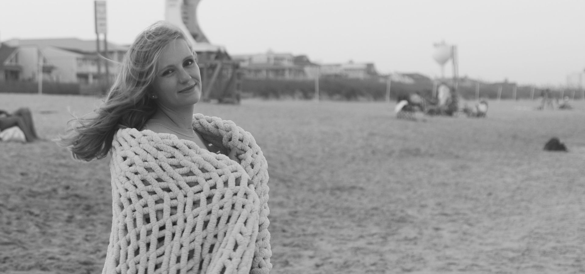A woman with long hair wrapped in a thick, knitted blanket, standing on a beach with a few people and structures in the background.