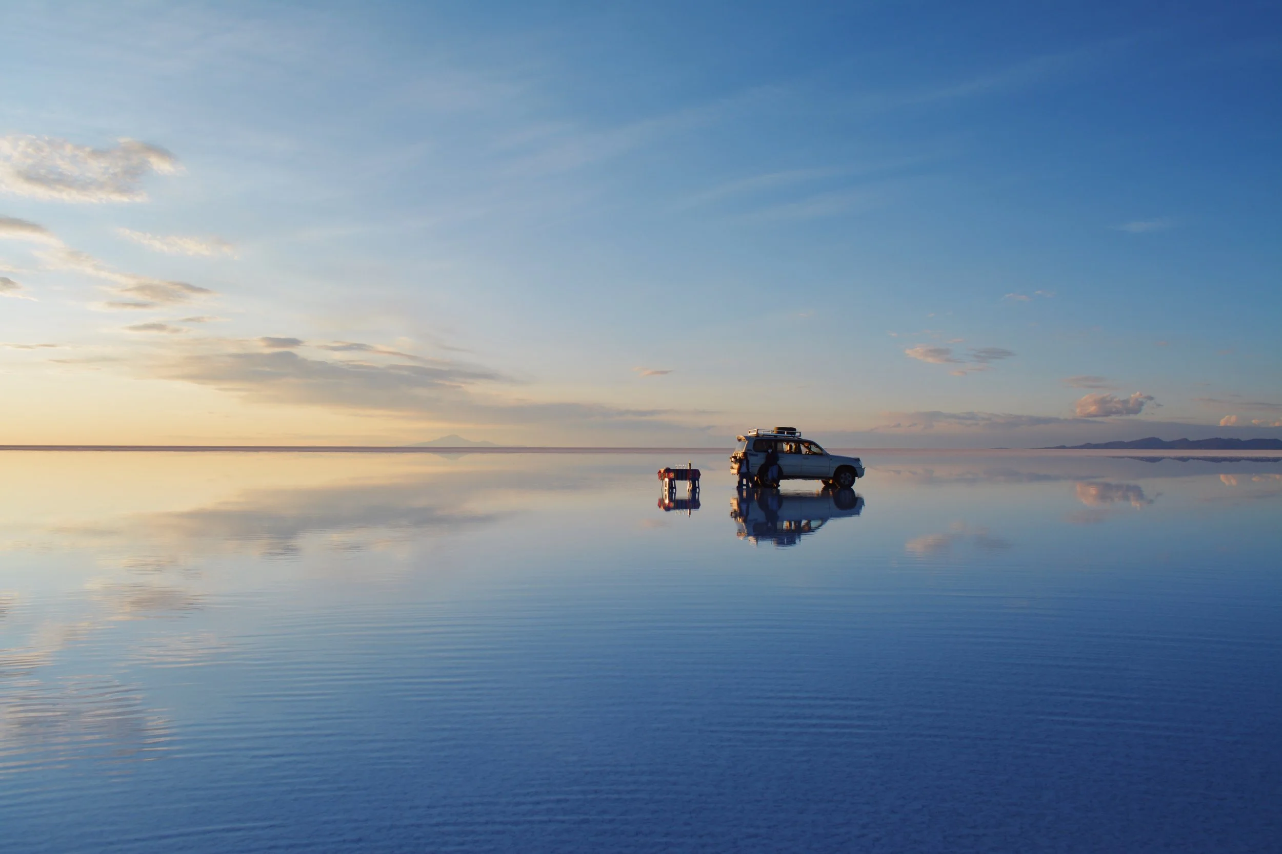 A vehicle parked on a vast, reflective salt flat at sunset with a small table and chairs beside it, creating a mirror-like effect on the ground with a colorful sky overhead.