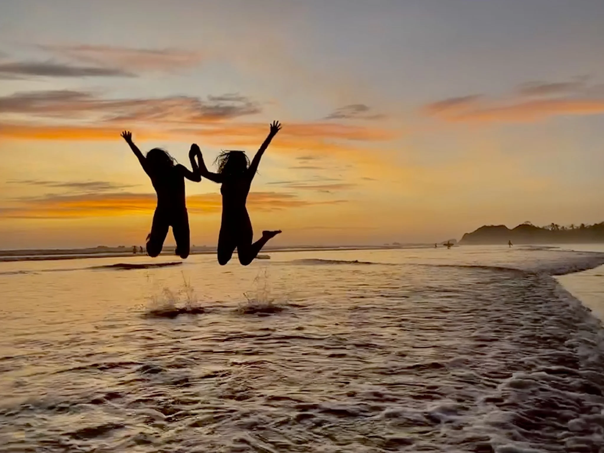 Two friends jumping together in the ocean beneath a golden sunset.