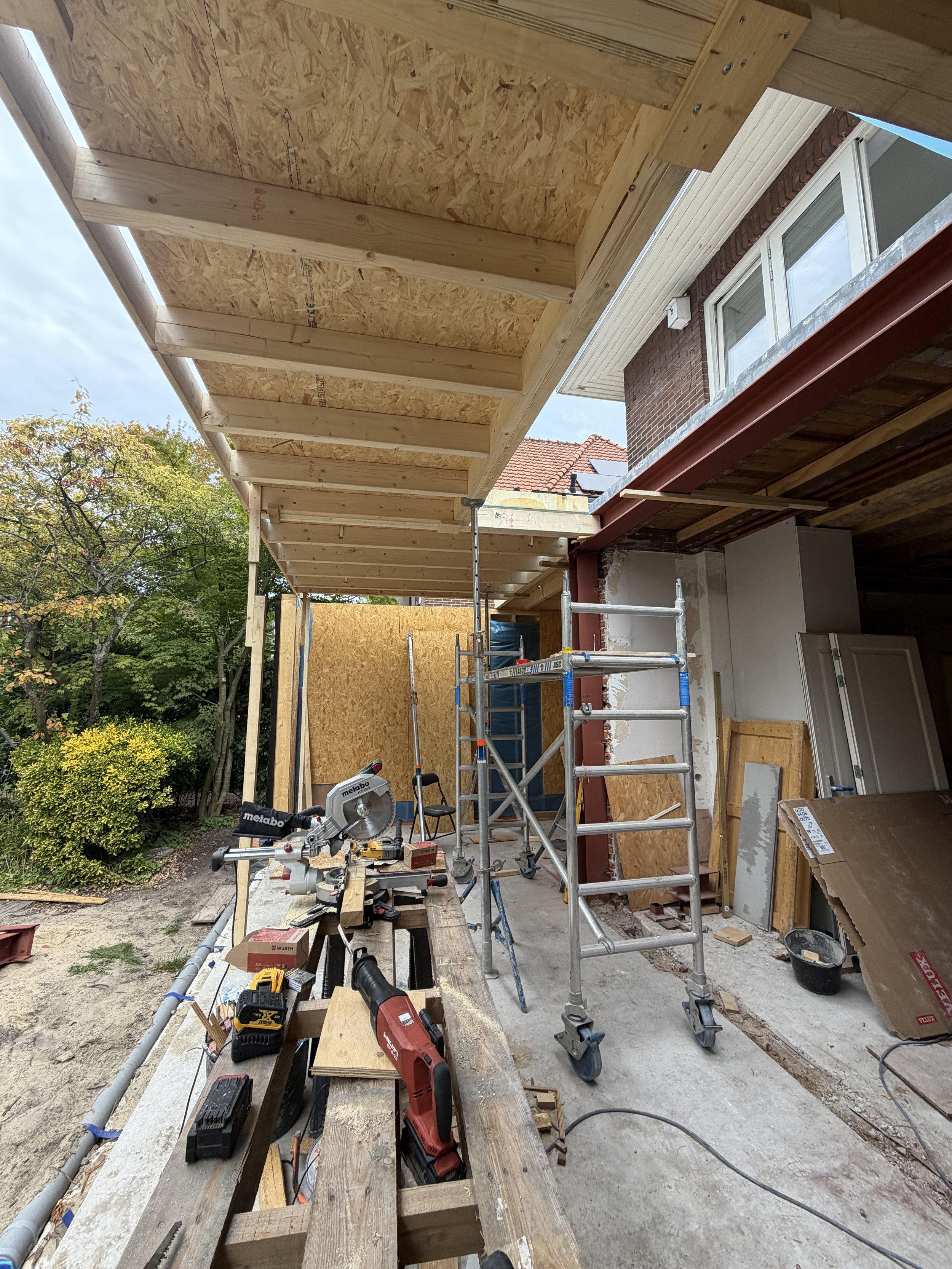 Bouwplaats met houten constructies en ladder, gereedschap en materialen voor de bouw van een overkapping, naast een huis met ramen en een tuin met bomen.