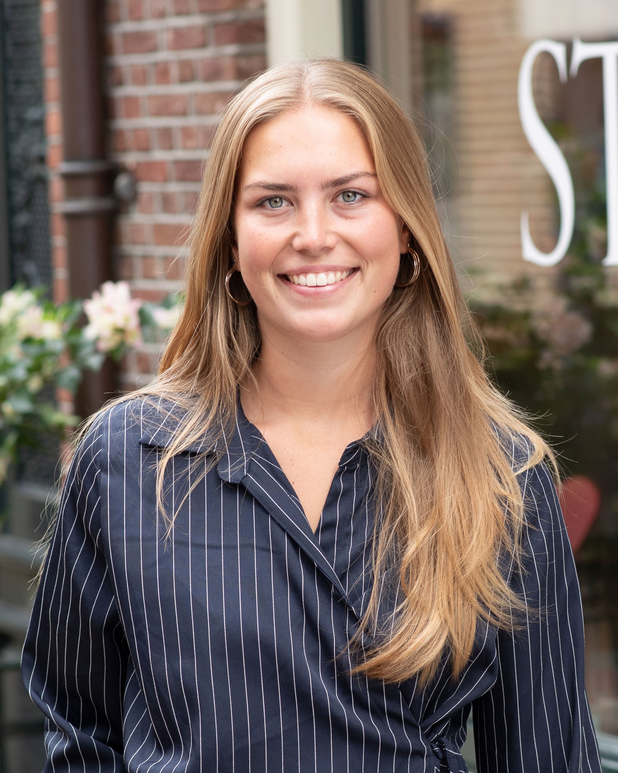 Close-up van een lachende vrouw met krullend haar, ronde glazen, dubbele ketting en oorringen, in een kantooromgeving.
