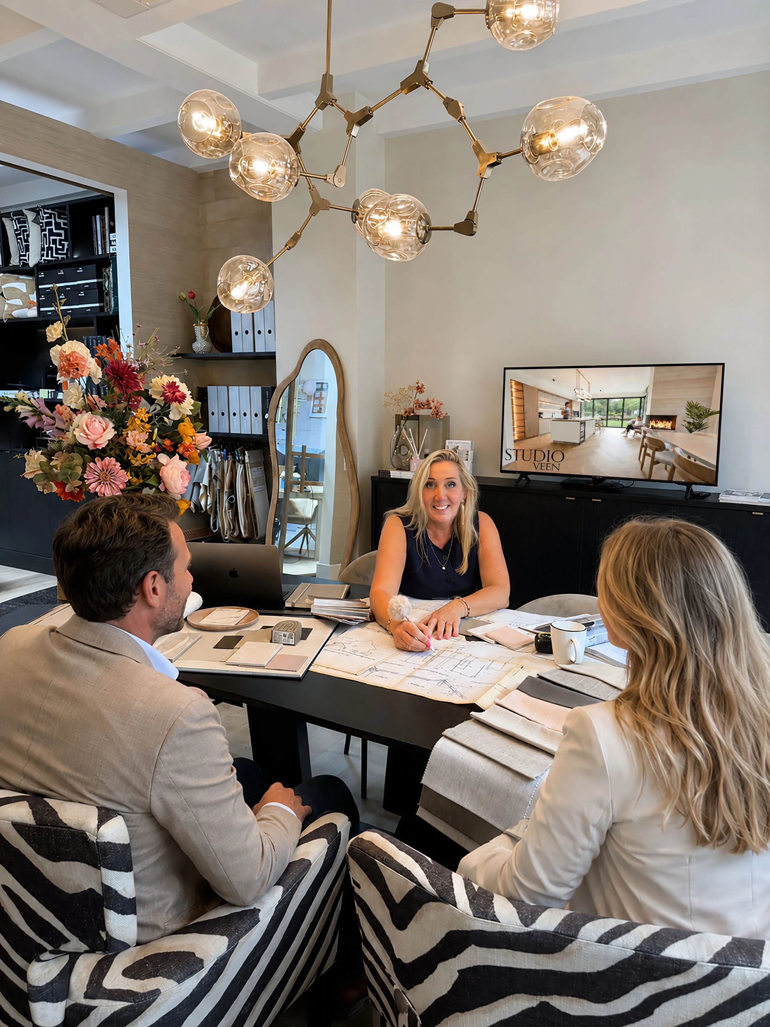 Vergadering van vijf mensen rond een tafel met documenten, laptops en kopjes in een modern interieur met een opvallende lichtarmatuur en een grote spiegel. In de achtergrond een tv-scherm en bloemen.