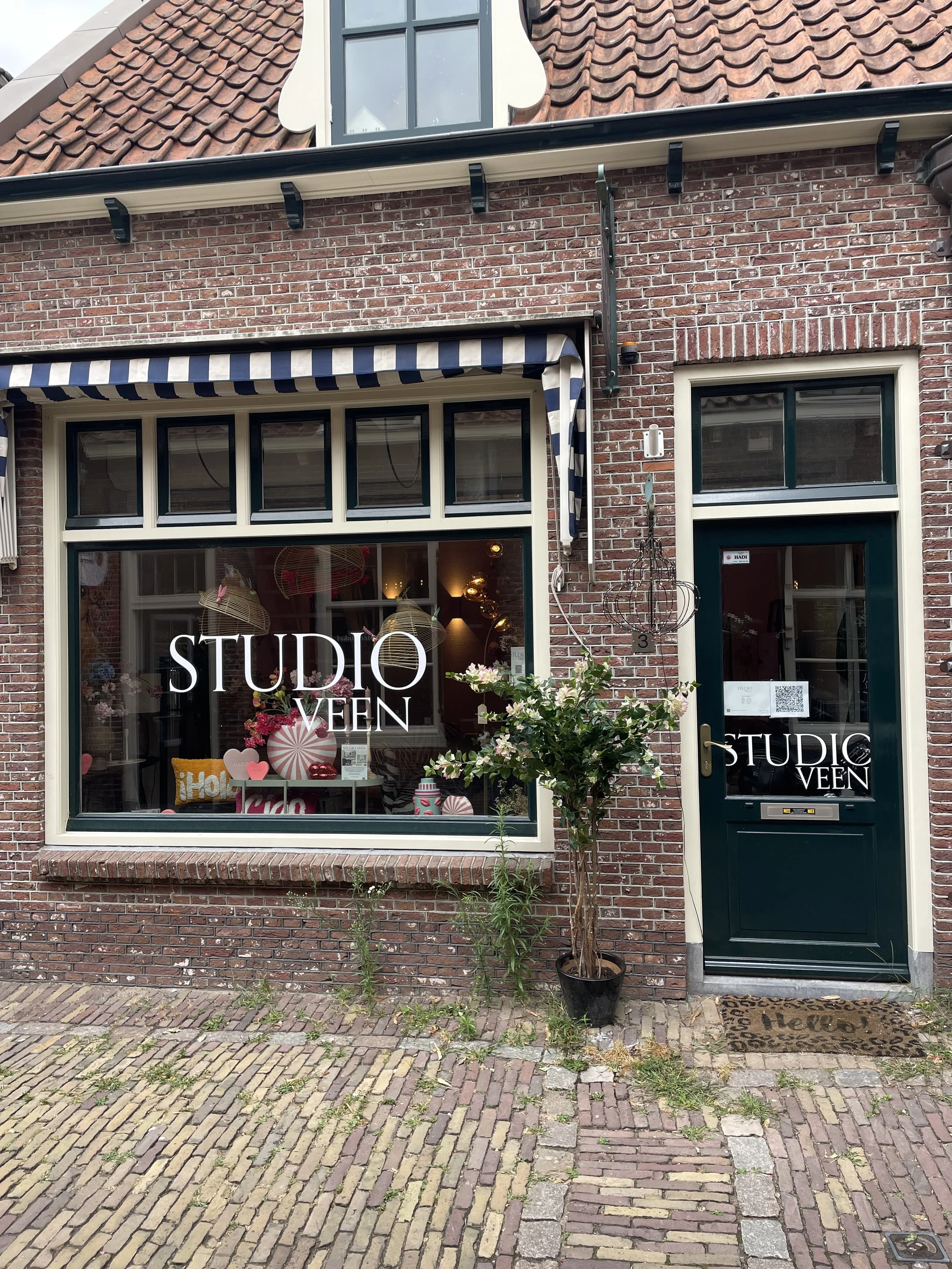 De gevel van een winkel met een grote raam en een deur, beide met de tekst 'STUDIO VEEN'. De winkel heeft een grote ramenpartij met een stoffen uitvalscherm met blauwe en witte strepen. Buiten staan planten, onder andere een grote pot met een boom. Het trottoir bestaat uit bakstenen. Boven de winkel bevindt zich een rood tegel dak en een klein uitbouw met een raam.