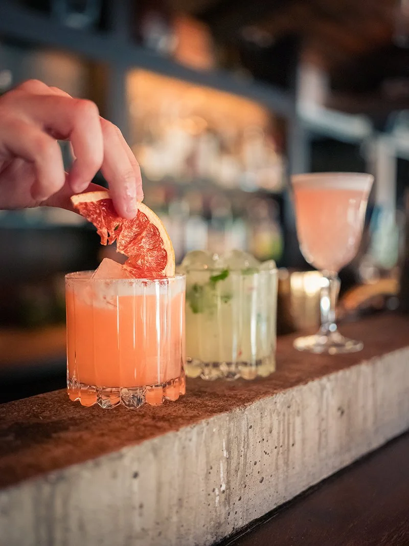 En hand som placerar en skiva blodorange i en rosa cocktail, med två andra cocktails i bakgrunden på en bar.