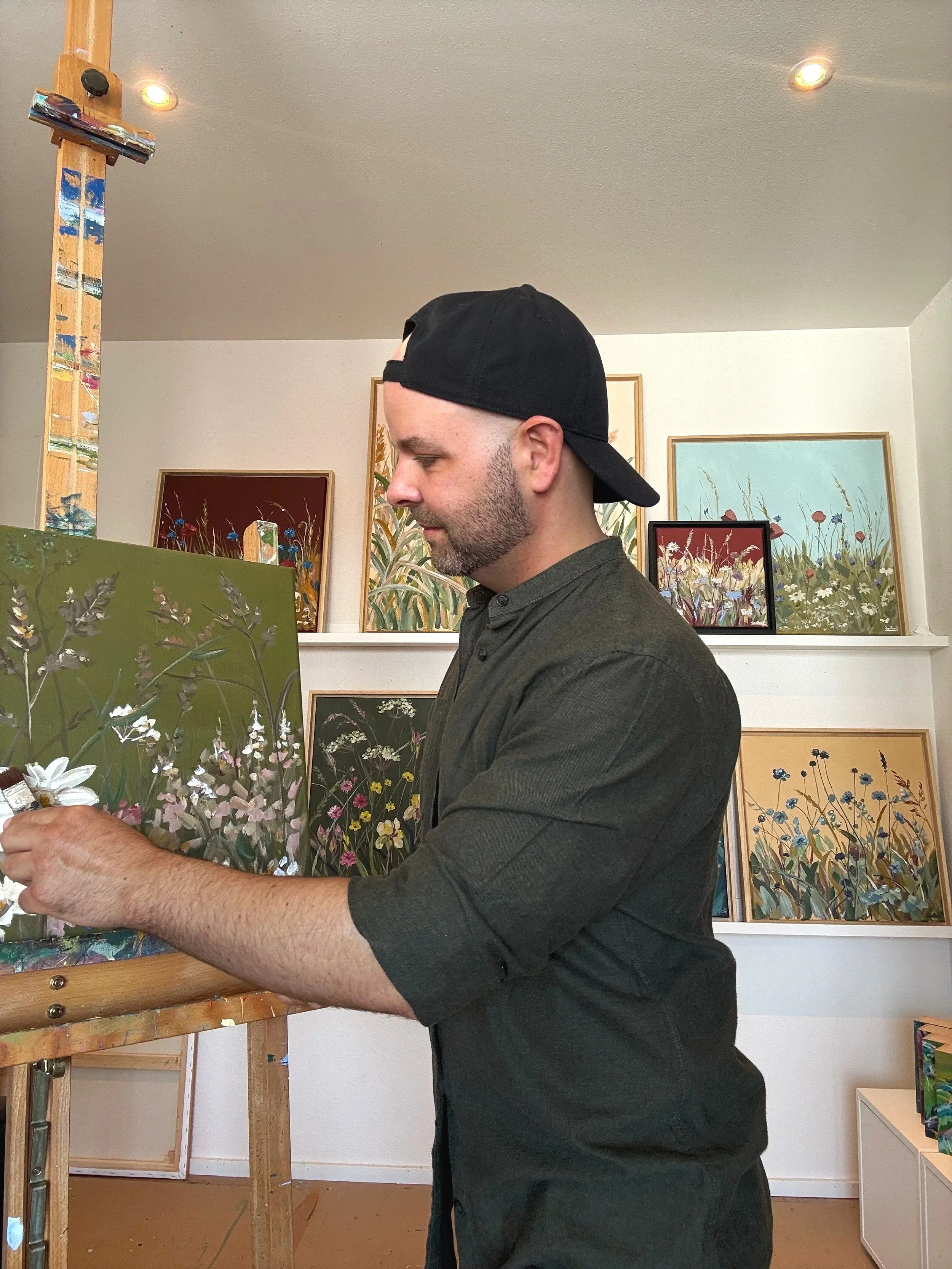 Ein Mann mit schwarzer Mütze, Bart und grauem Hemd arbeitet in einer Kunstgalerie, umgeben von mehreren Gemälden mit Blumen- und Pflanzenmotiven an der Wand.