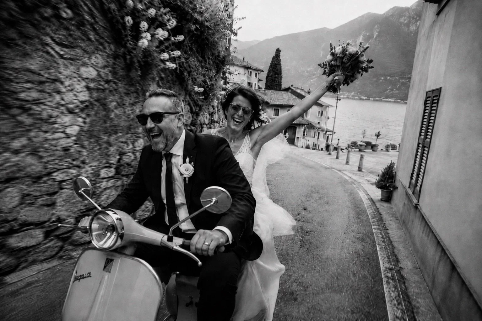 Bride and groom riding a vintage Vespa through a narrow Italian village street by the lake, joyful documentary-style wedding photography with cinematic black and white atmosphere and natural movement.