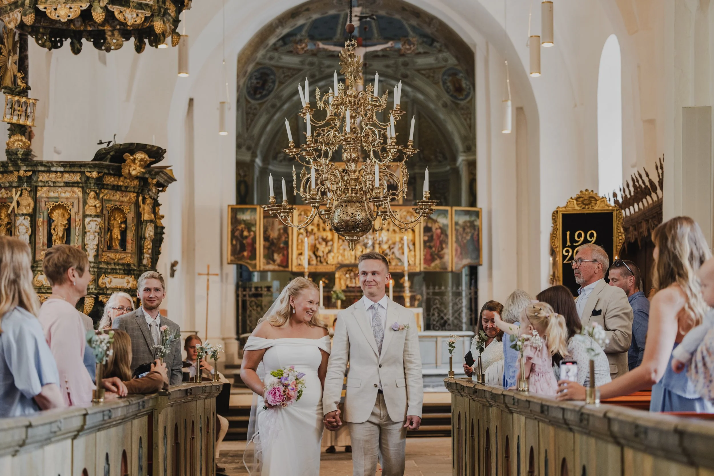 A wedding ceremony inside a church, with a bride and groom holding hands and walking down the aisle, surrounded by guests. The church features a large chandelier, ornate gold decorations, and religious artwork in the background.