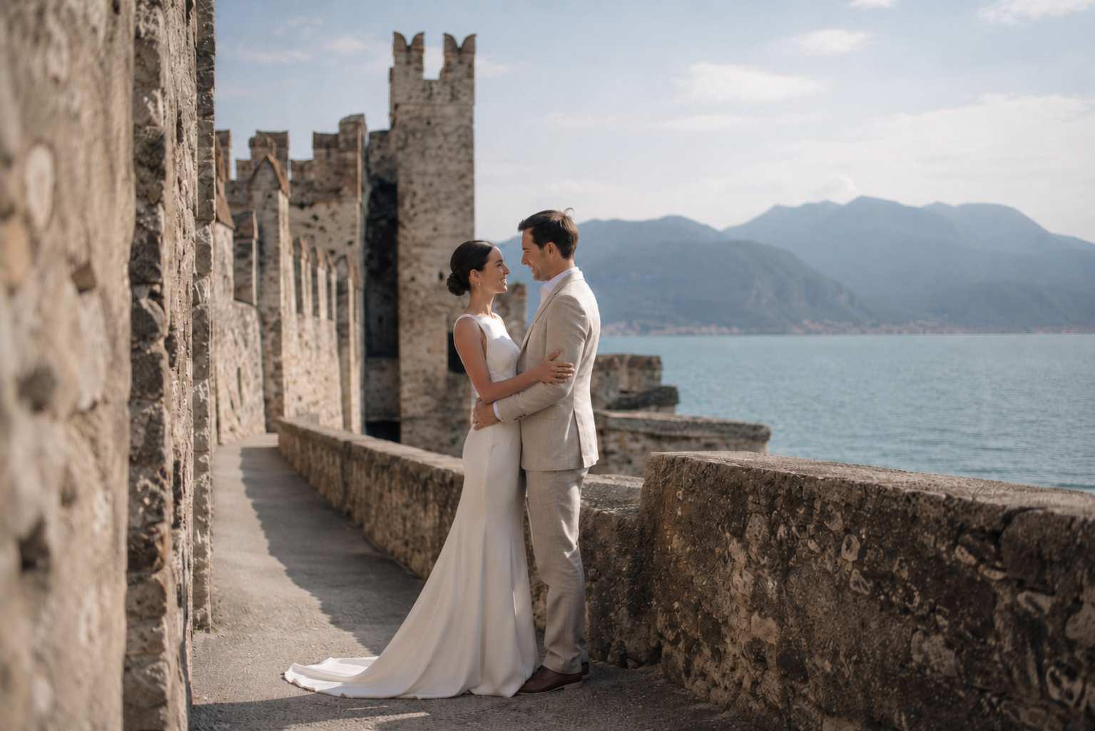 Romantic civil wedding portrait at a historic Lake Garda castle overlooking the lake and mountains during golden hour. Bride and groom embracing naturally with soft editorial light and an intimate documentary feel.