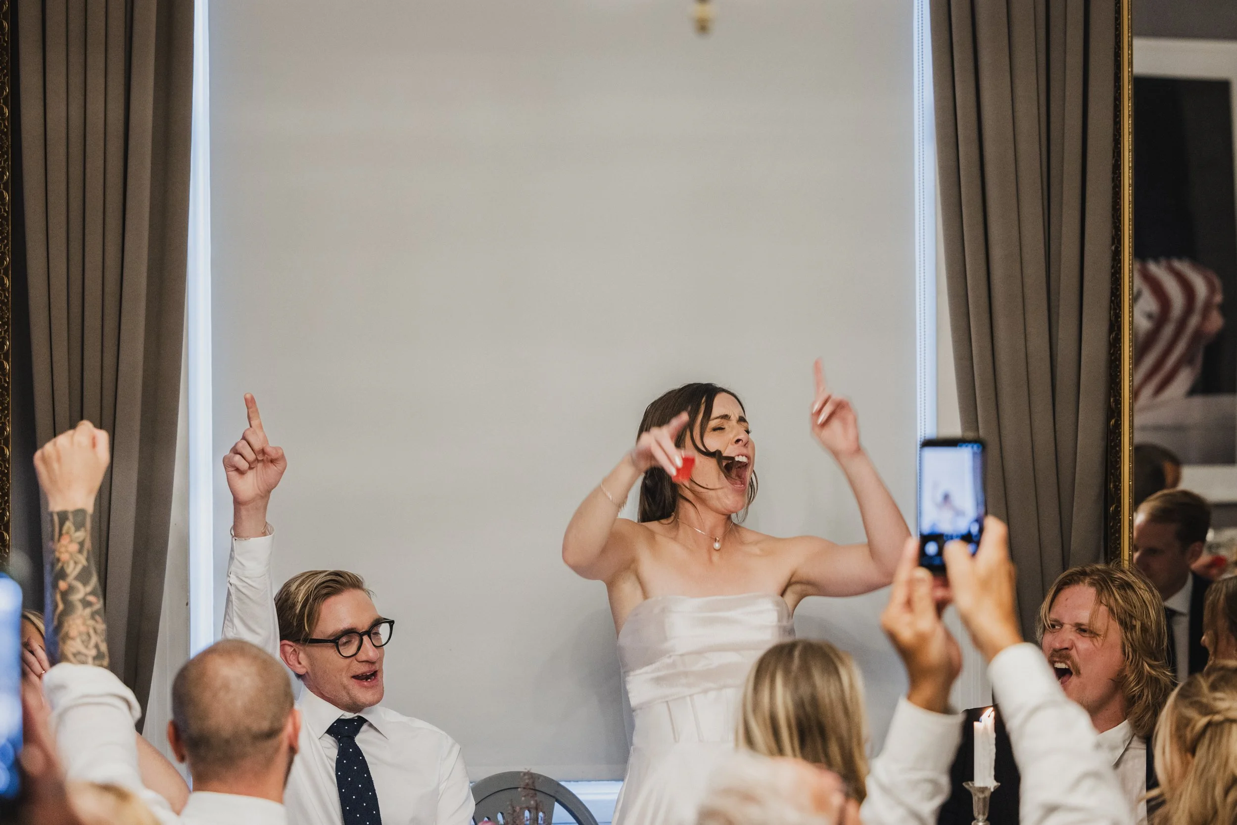 A woman in a strapless wedding dress celebrating with raised arms while surrounded by wedding guests at a reception.