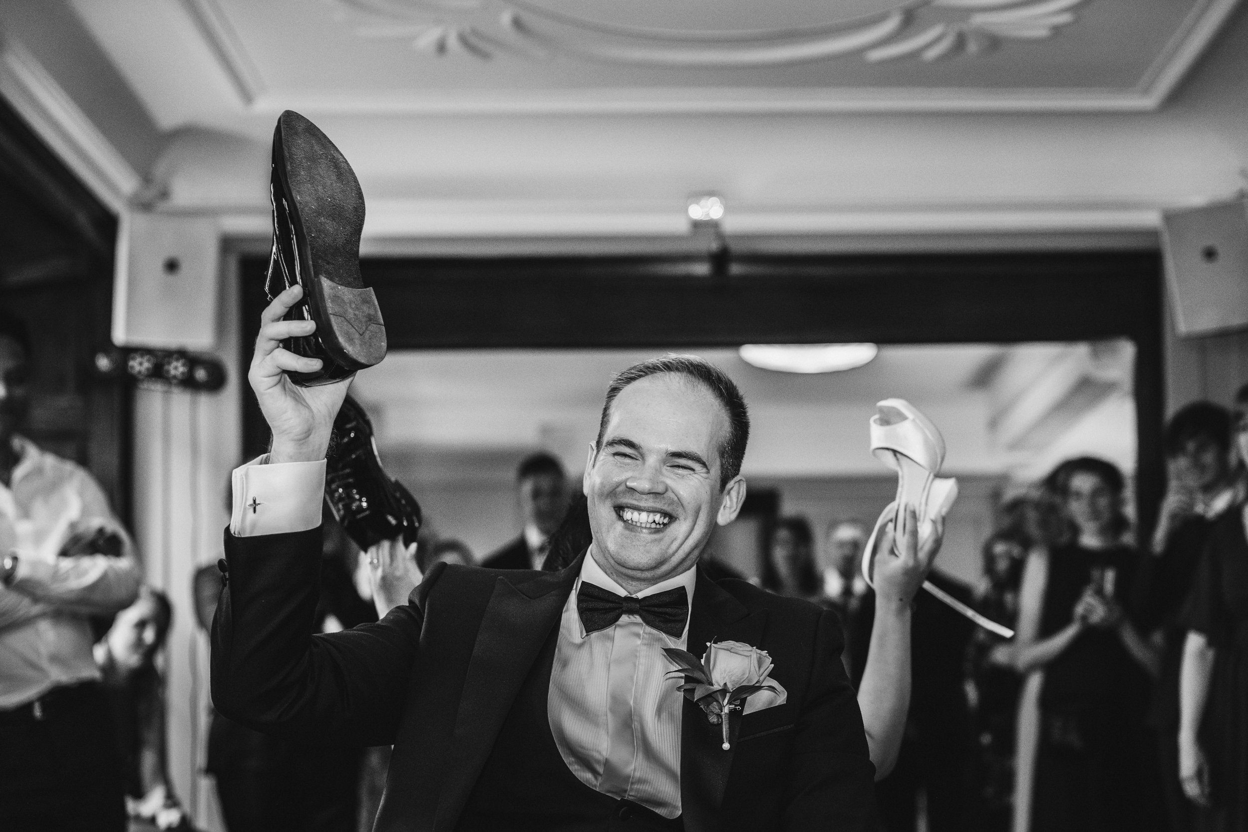 A man in a tuxedo and bowtie smiling and holding up a shoe in each hand at a celebration or wedding, with people in the background.