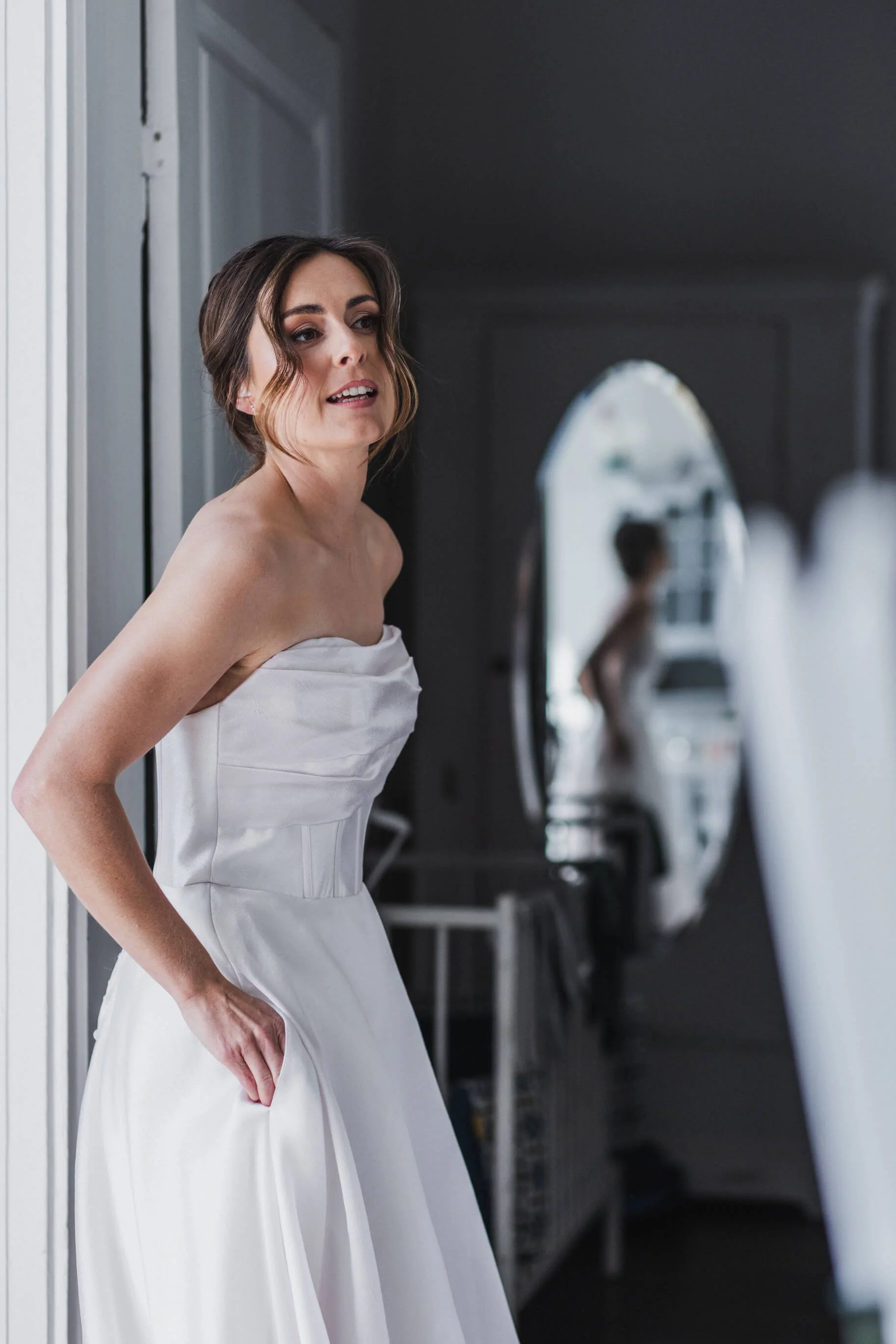 En ung kvinna i en vit brudklänning står vid en dörr, tittar ut genom fönstret, och reflekteras i en oval spegel i bakgrunden.