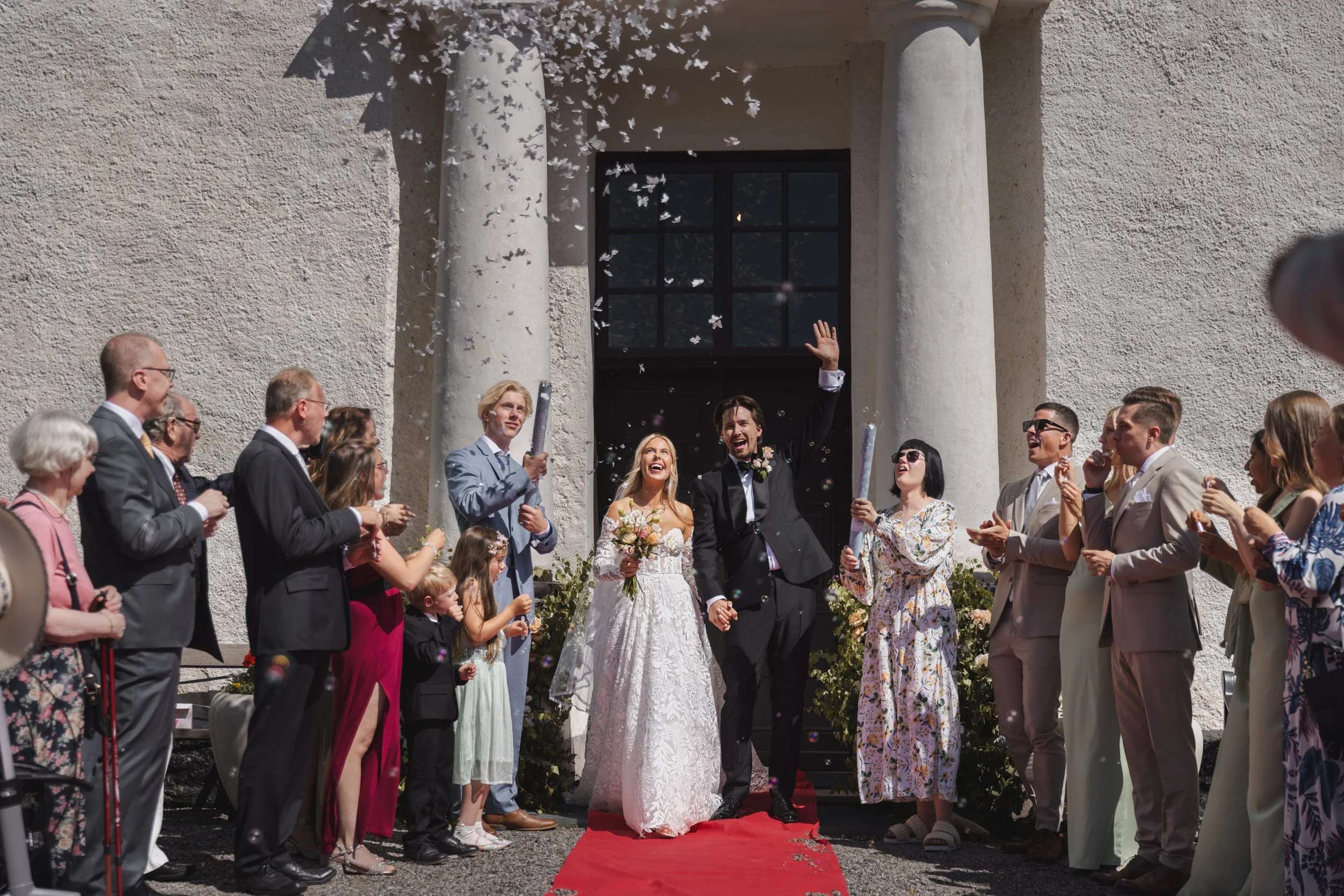 En brud och brudgum som går längs en röd matta utanför en kyrkobyggnad, omgivna av gäster som kastar konfetti.