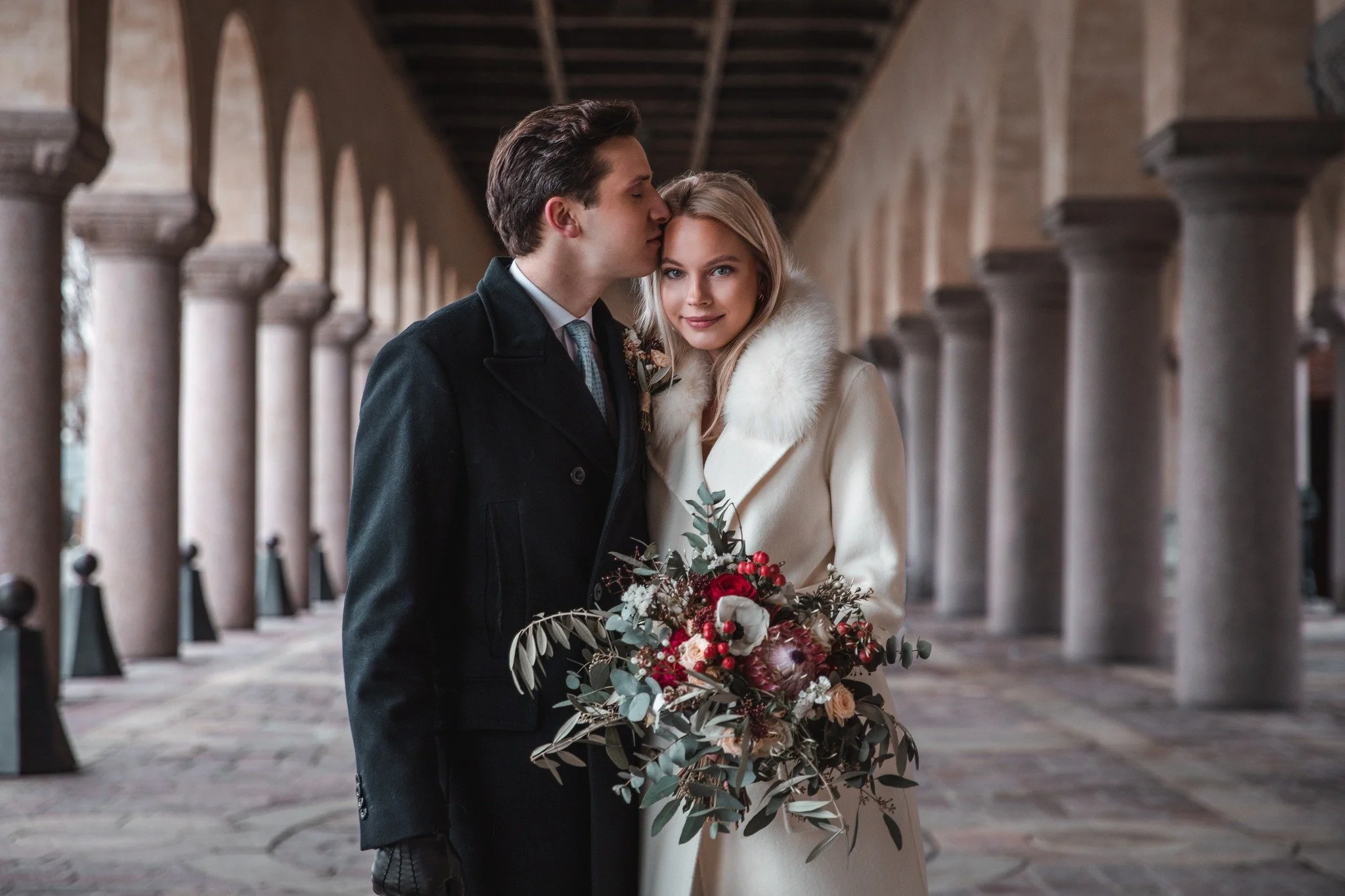 A couple on their wedding day standing in a colonnade. The groom is kissing the bride on the head, and she is holding a bouquet of flowers. The bride is wearing a white coat with a fur collar, and the groom is in a dark coat.