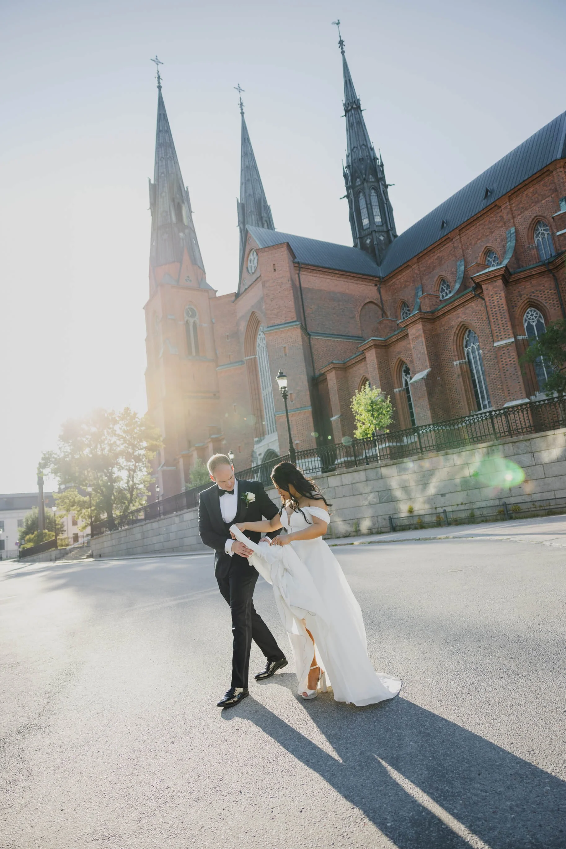 En brud och brudgummen går tillsammans på en öppen gata framför en stor, äldre kristen kyrka, med solen som lyser starkt bakom dem och kastar långa skuggor.