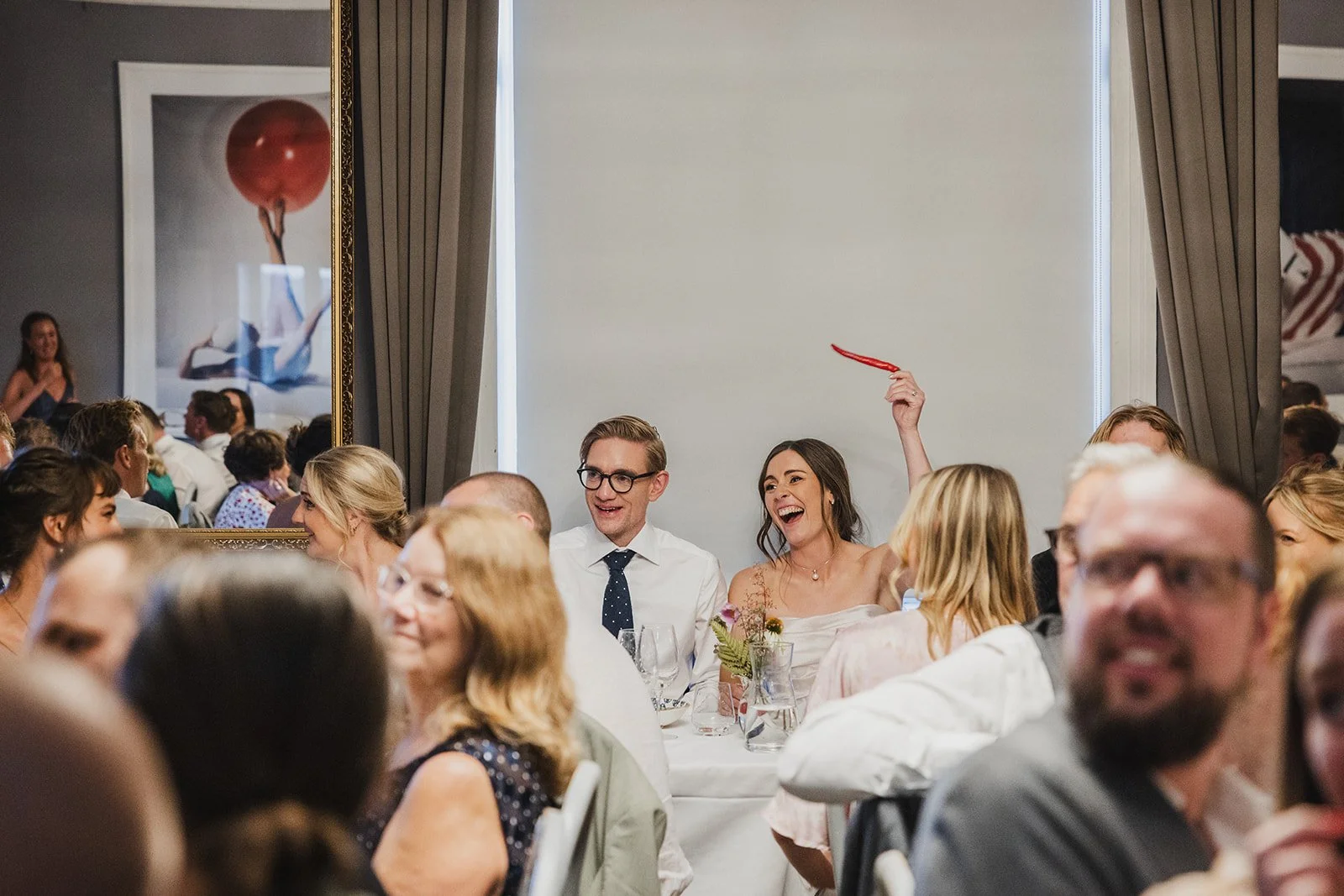 A wedding reception with guests seated at tables, a woman raising a red object, laughing among the guests, and a large mirror reflecting part of the room.