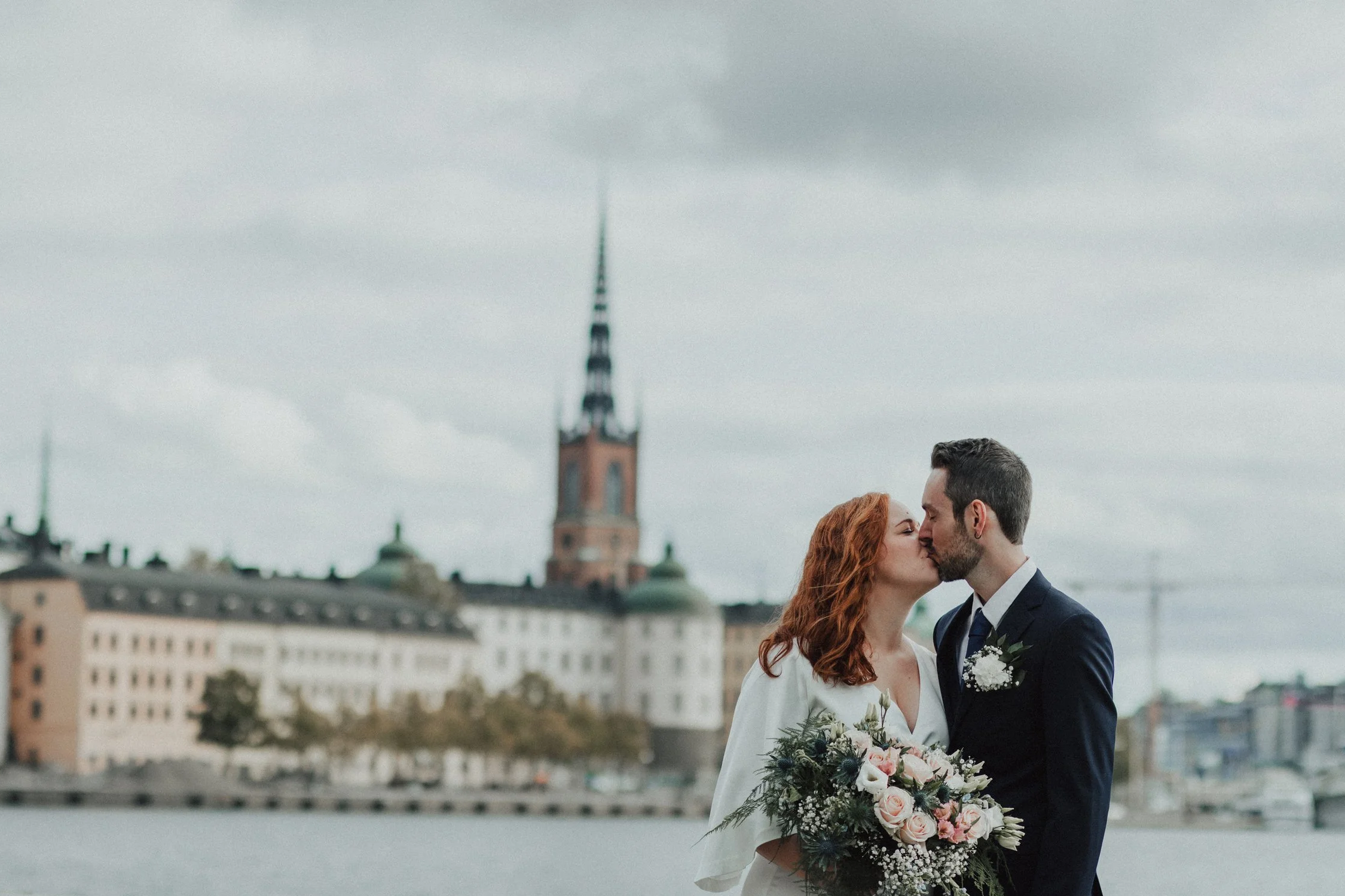 A bride and groom kiss outdoors during their wedding, with a cityscape and church steeple in the background.