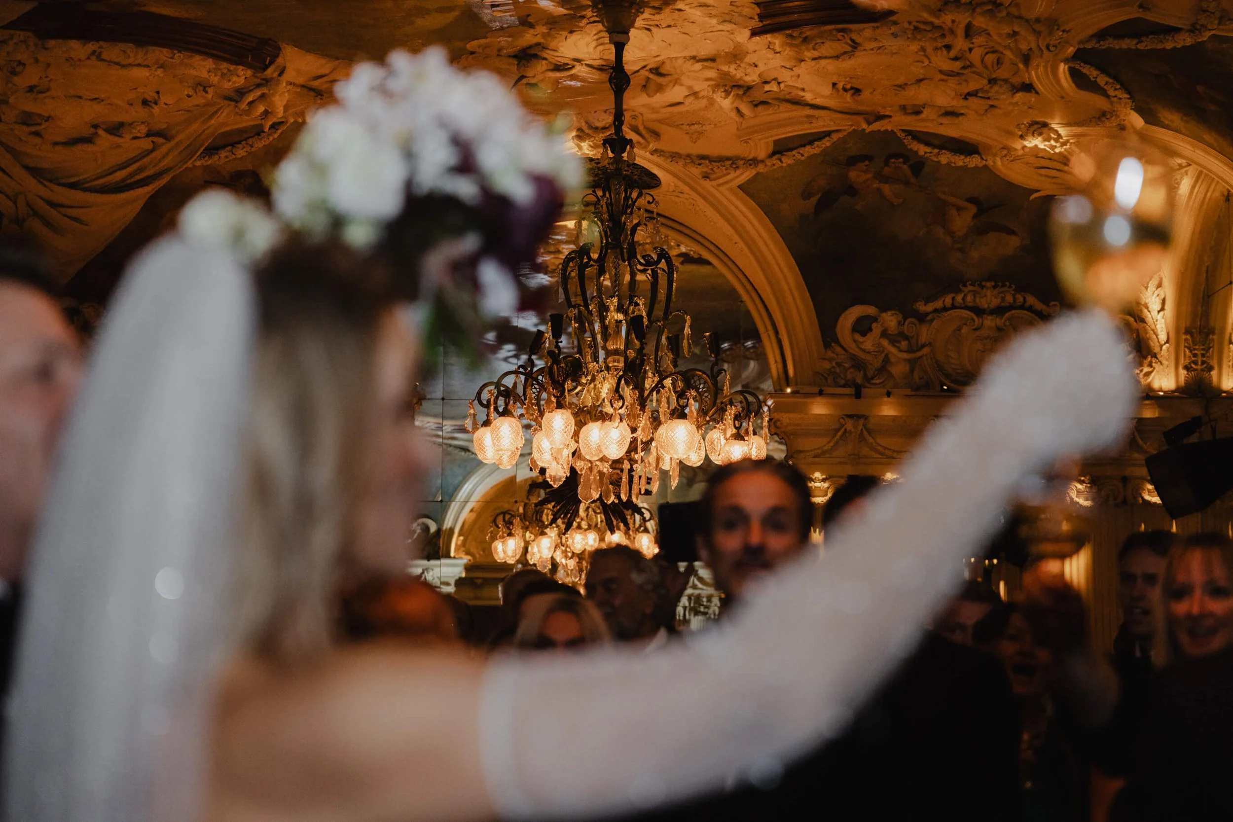 Bride entering Café Opera beneath historic chandeliers as guests welcome the couple