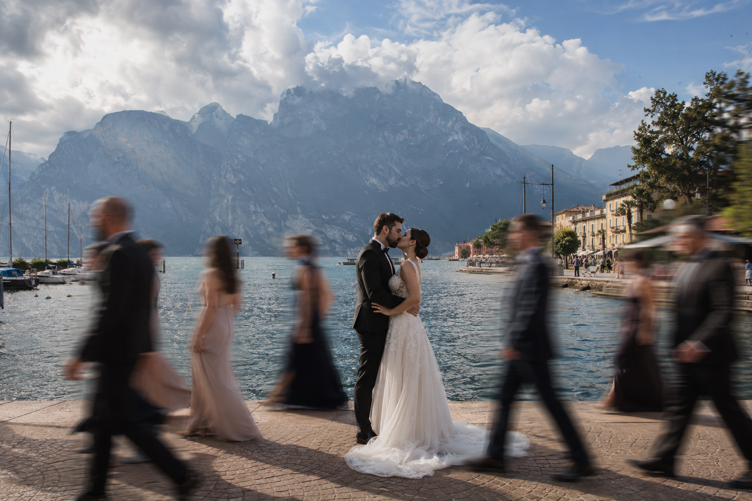 Bride and groom embracing on the lakeside of Riva del Garda with dramatic mountain scenery, surrounded by bridal party walking in soft motion blur, creating a cinematic and documentary-style wedding moment with natural movement and Lake Garda feeling