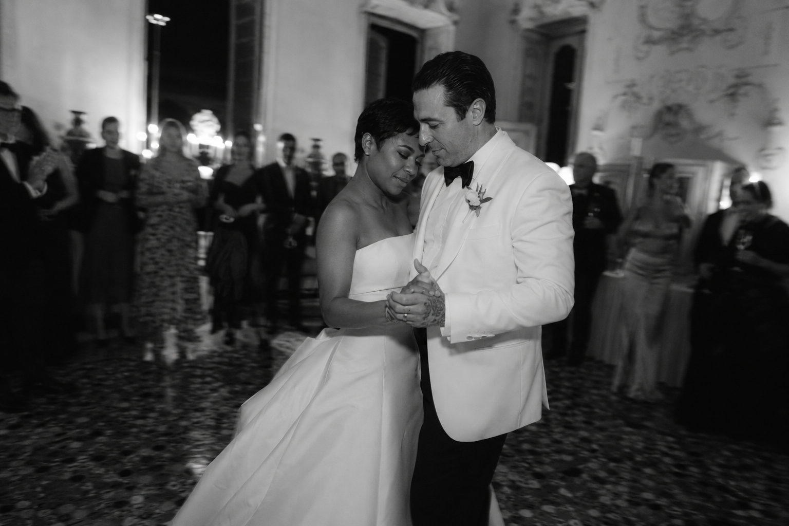 Black and white photo of a couple sharing their first dance inside Villa Sola Cabiati during an intimate Lake Como wedding reception, surrounded by guests and candlelit atmosphere.