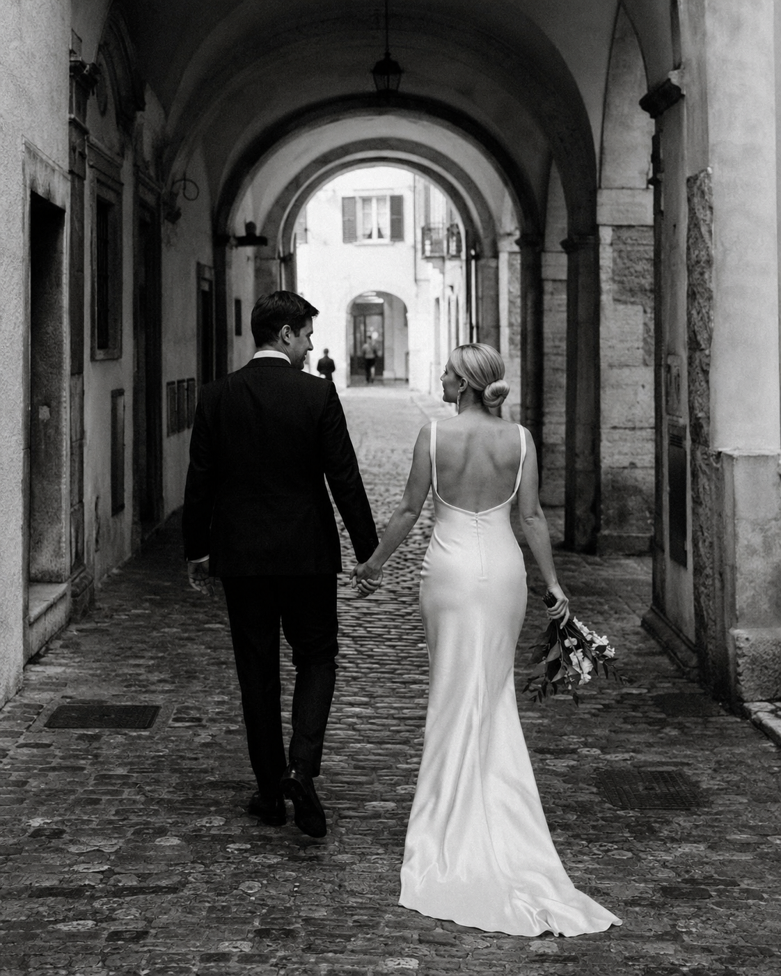 Black and white Trento wedding photo of a bride and groom walking through historic stone arches in Trentino, Northern Italy