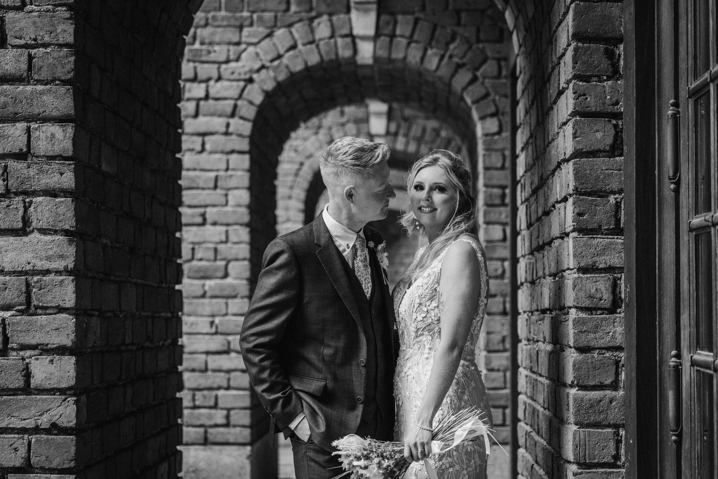 A black and white photograph of a newlywed couple standing close together in a brick archway. The groom is wearing a dark suit and tie, looking at the bride. The bride is holding a bouquet and smiling at the camera, wearing a lace wedding dress.