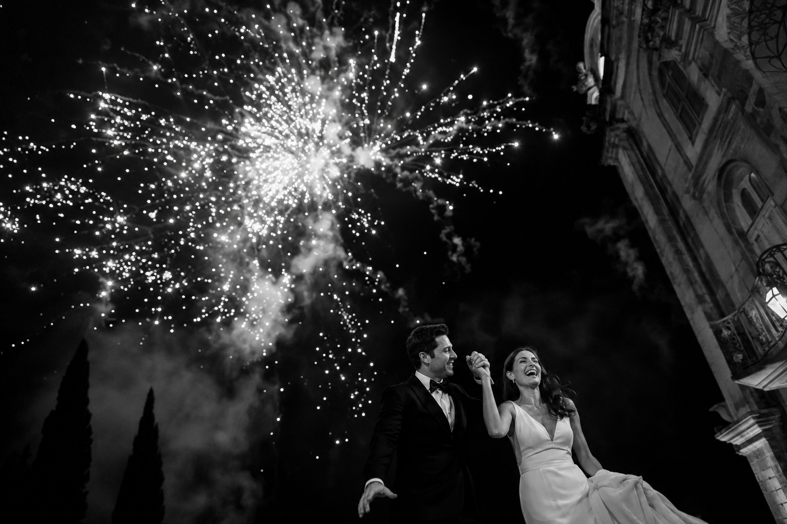 Bride and groom celebrating beneath fireworks during an elegant nighttime wedding in Tuscany, Italy, cinematic black and white documentary photography with joyful movement and dramatic atmosphere.