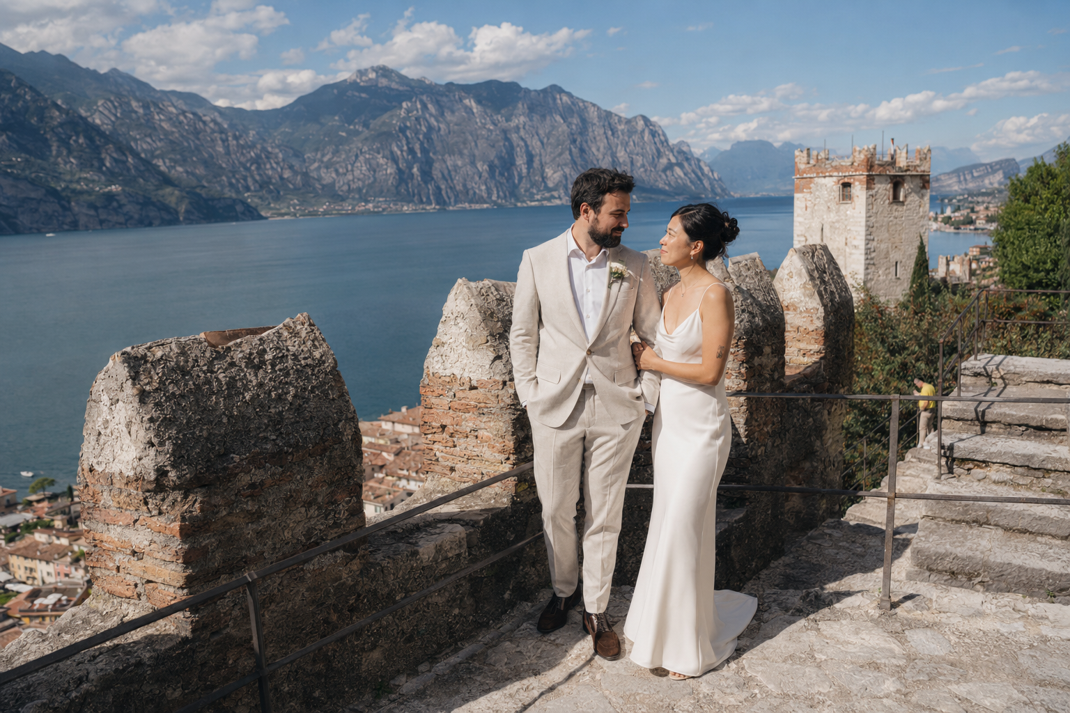 Alt text:  Bride and groom standing on the medieval walls of Castello Scaligero di Malcesine overlooking Lake Garda and the surrounding mountains during a destination wedding in Italy.