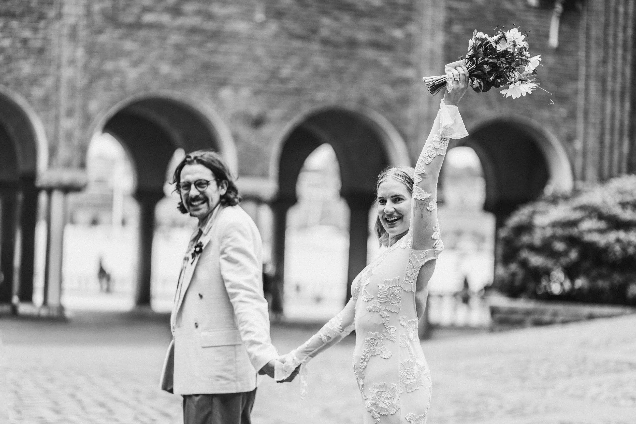 A black and white photo of a smiling couple holding hands outdoors in Stockholm's Stadshuset. The woman is holding a bouquet of flowers with her left arm raised. The background features arched brick structures and some greenery.
