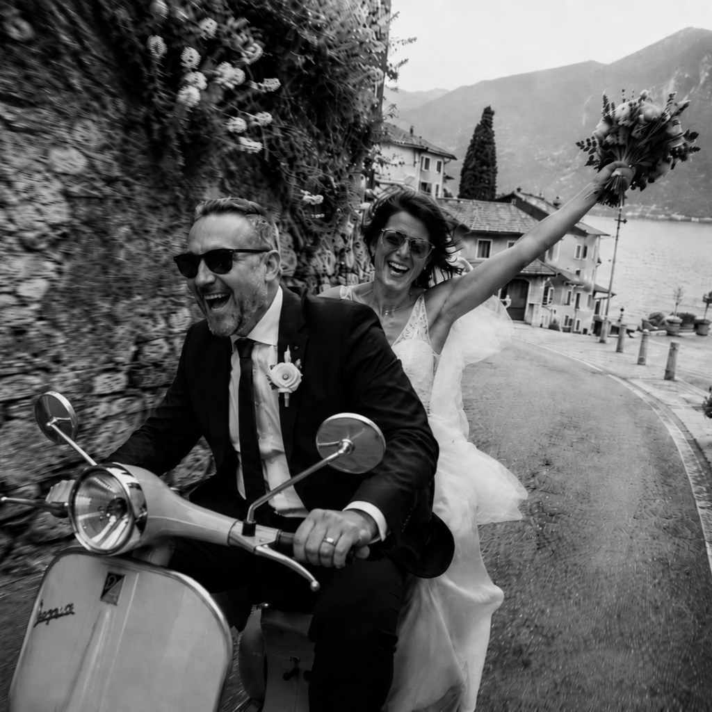Romantic black and white wedding photo of a bride and groom riding a Vespa through a narrow Italian lakeside village street near Lake Garda, with mountains and lake views in the background.