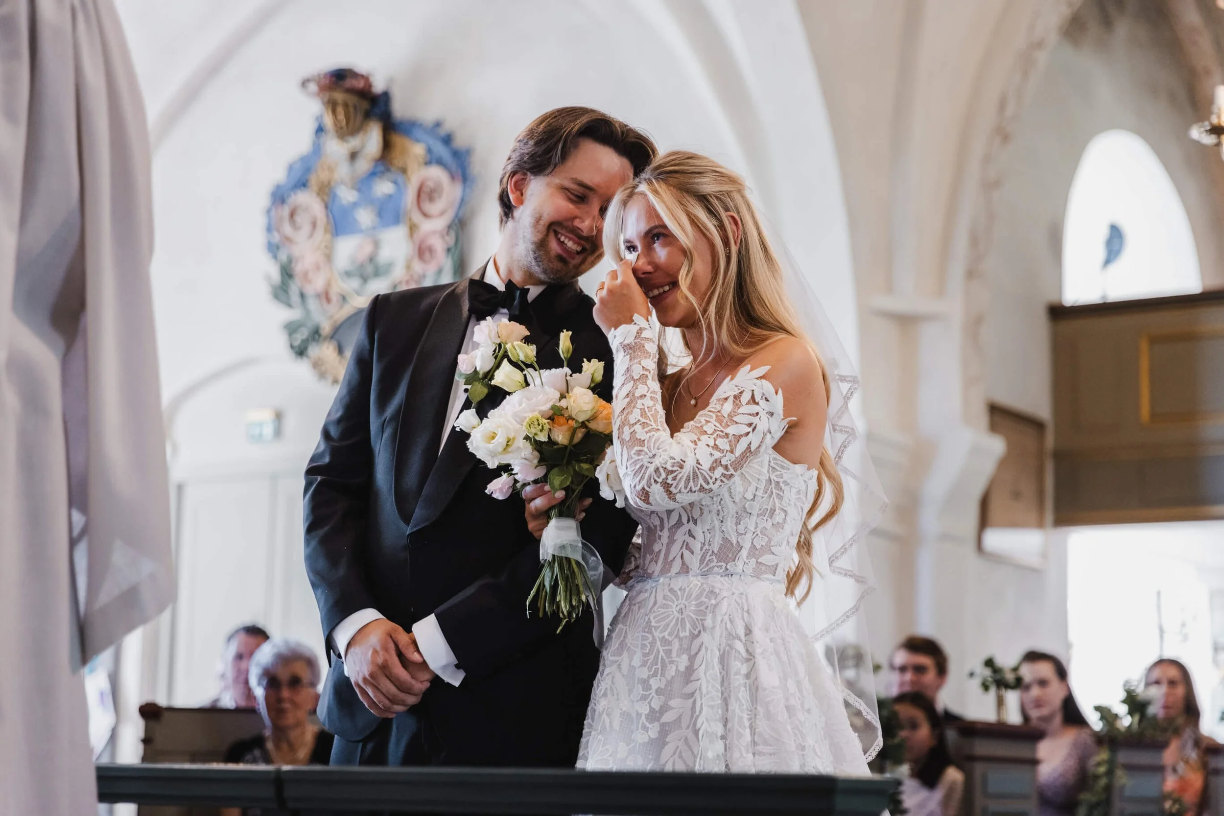 En brud och en brudgrum i svart smoking som håller en blombukett under ett bröllop i en kyrka, med gäster i bakgrunden.