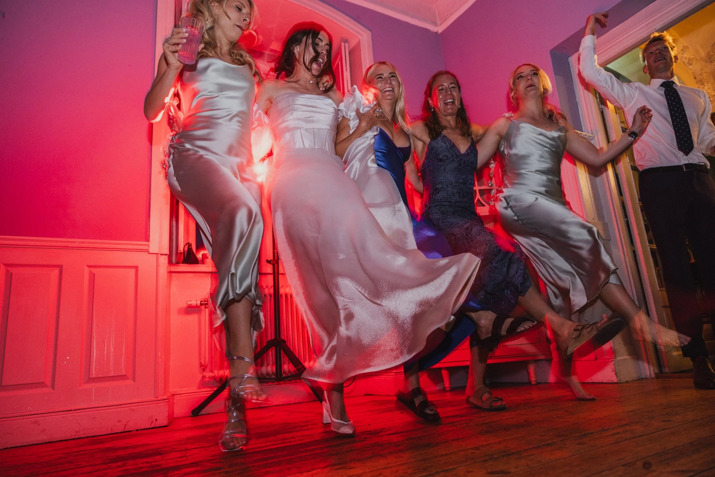 Group of women in dresses and one man in a shirt and tie dancing or celebrating at a party or wedding reception with pink and yellow lighting.